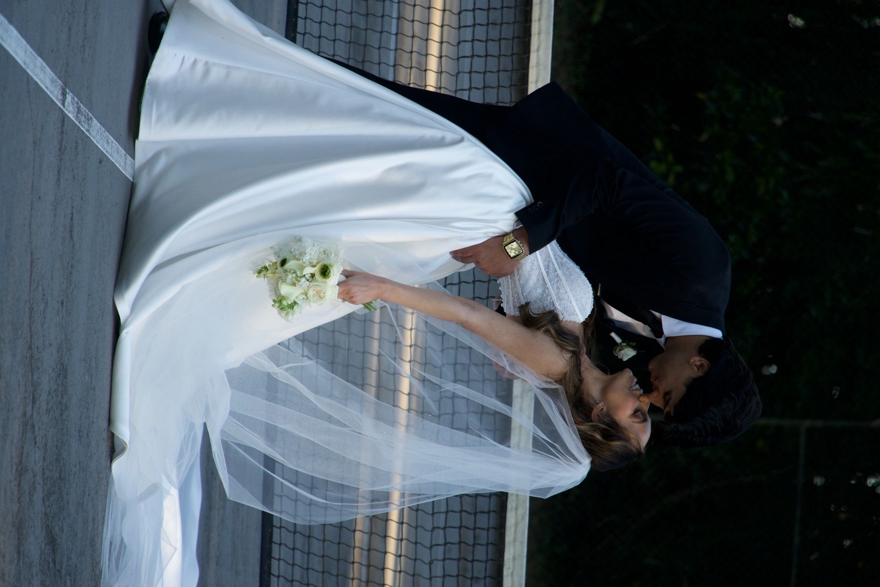 A groom dips his bride into a kiss on a tennis court, her veil trailing across the ground