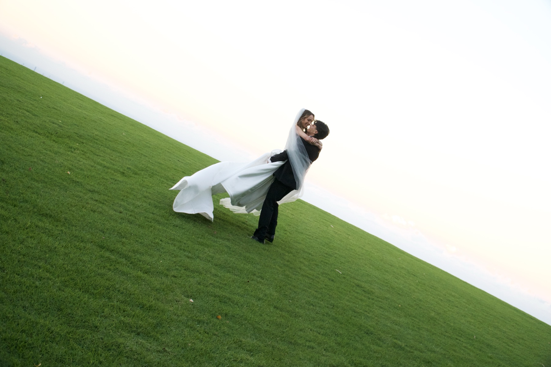 A groom lifts his bride on an open grass hill against a pale evening sky