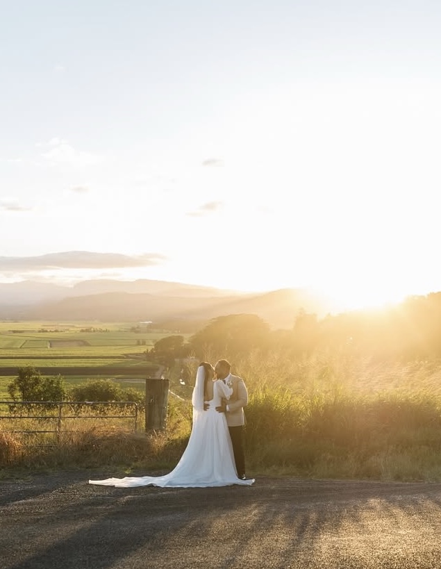 A bride and groom kiss at a farm gate, the sun setting low over the valley behind them