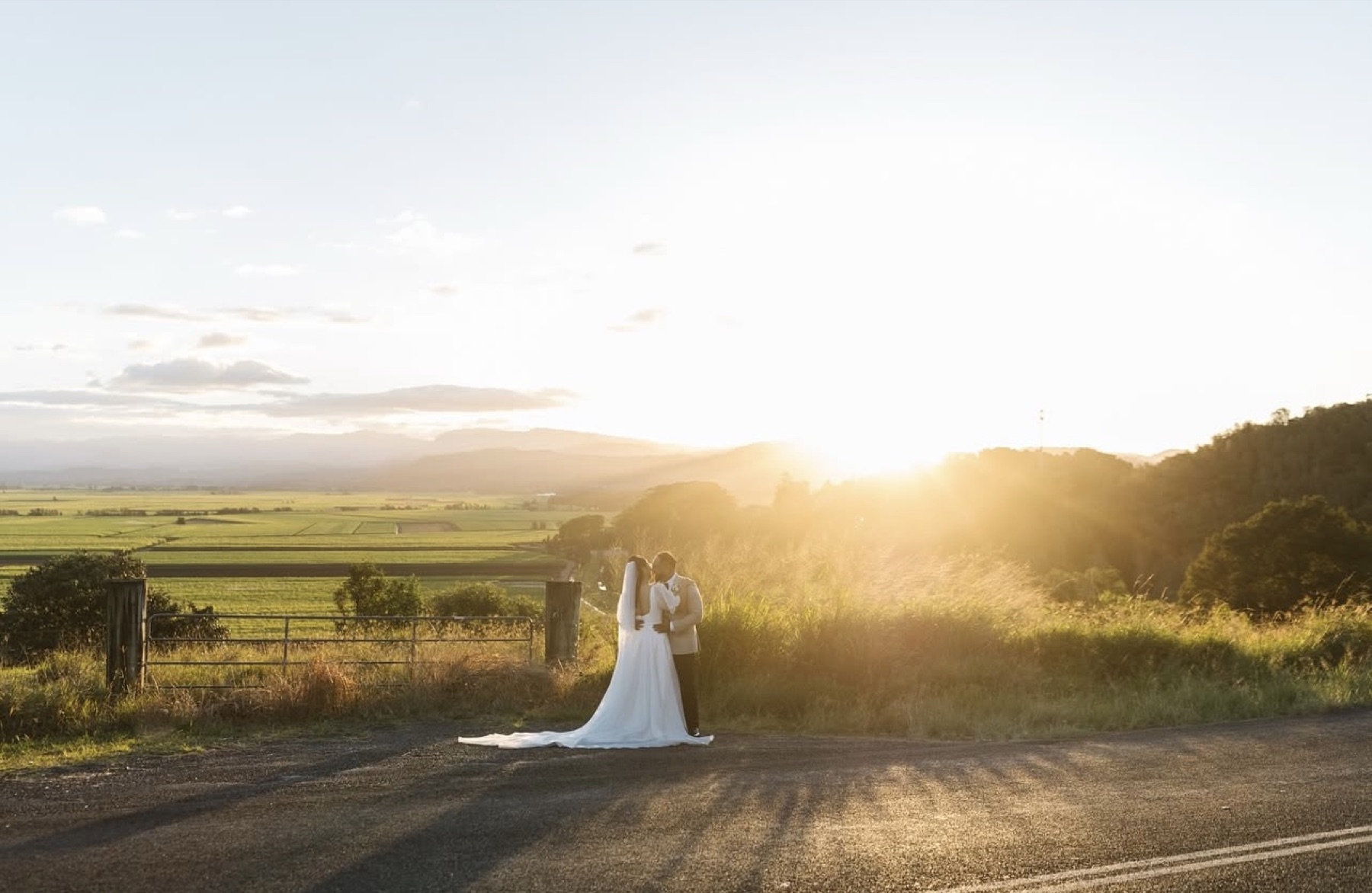 A bride and groom kiss at a farm gate, the valley and ranges gold behind them
