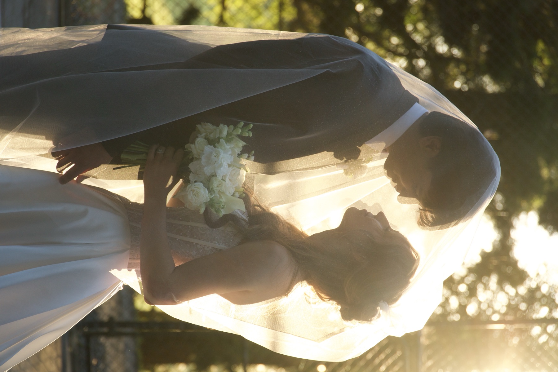 A couple stand forehead to forehead beneath a veil lit from behind by low sun
